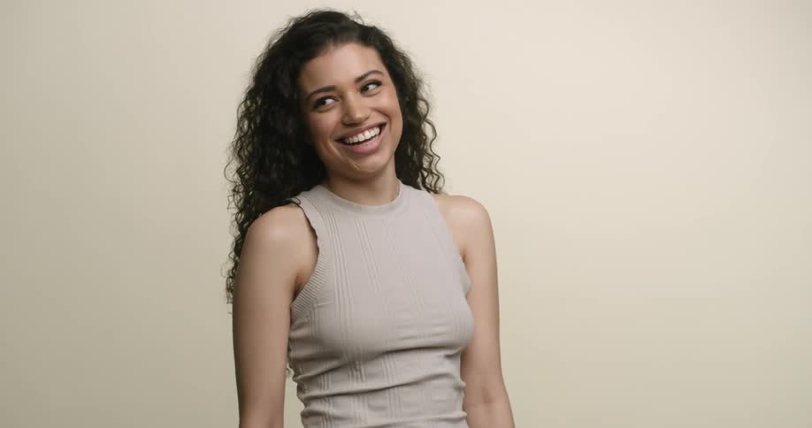 A young, ethnic female with dark, curly hair, beautiful teeth, and full lips gives a beaming smile and laughs at something behind the camera.