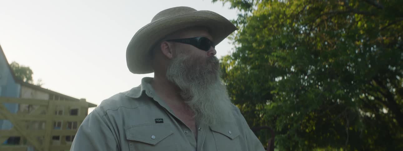 A man with sunglasses and a hat eating with trees the sky and clouds in the background