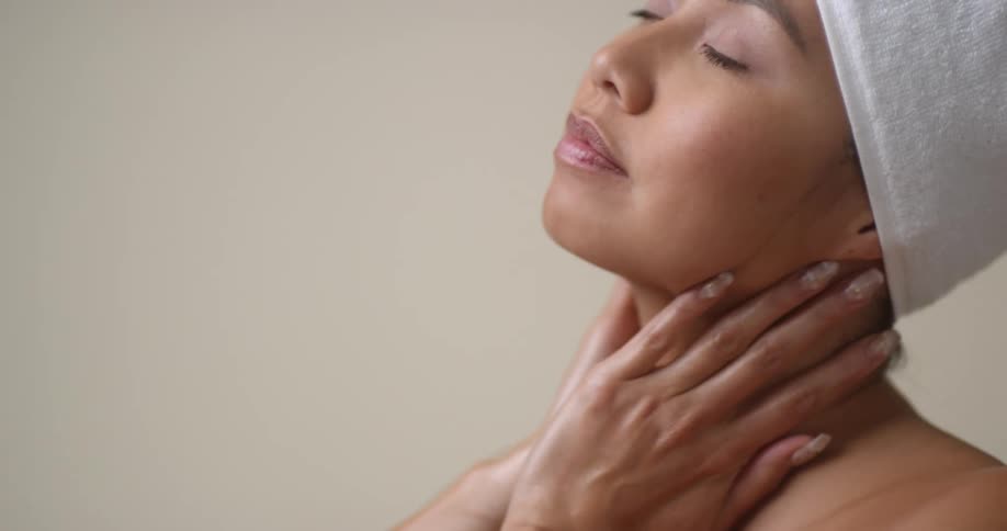 An Asian woman with a towel on her head gives a big yawn, smiles, stretches, and then rubs her hands down her neck and chest.