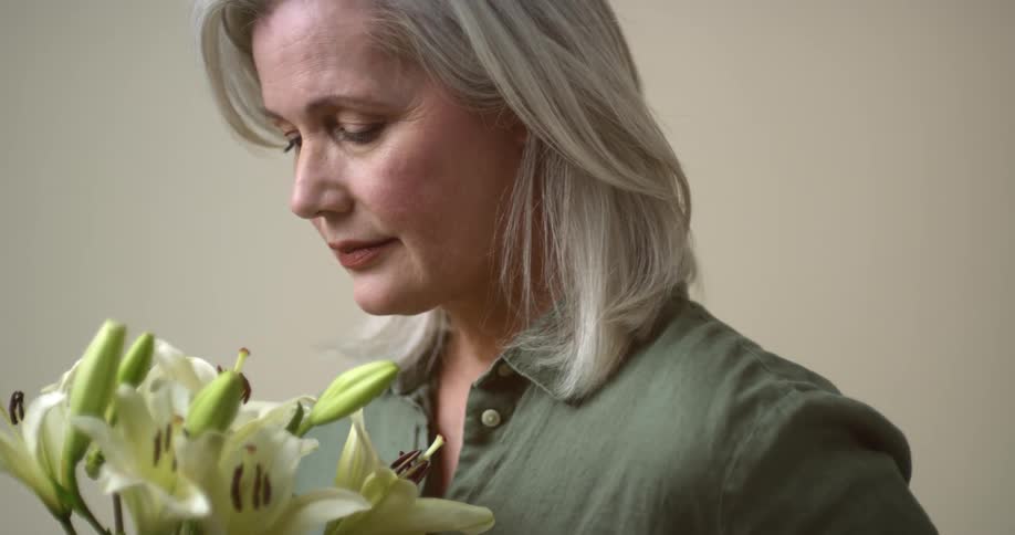 An older white woman with silver hair and a mature face holds a bunch of lilies and looks thoughtfully at them and then up into the distance.