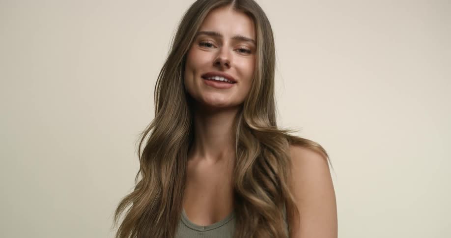 A young, white woman with light hair and eyes looks down at the camera over her shoulder and then moves to her other shoulder while giving a shy smile