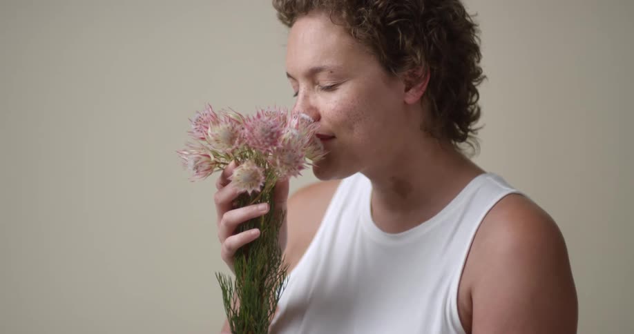 A young, Caucasian woman with boy-cut curly hair sniffs a bouquet of flowers and then turns to the camera with a big dimpled smile.