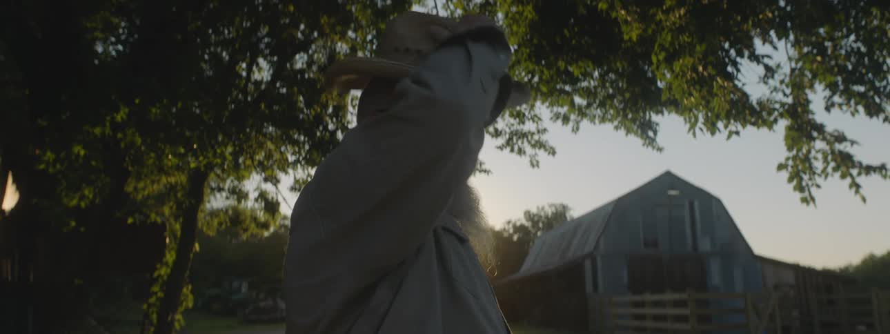A man standing on a farm removes his hat to wipe the sweat from his head before replacing it