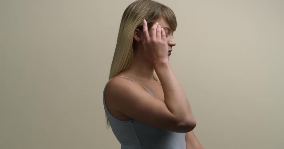 A young, blonde, Caucasian female in profile looks down shyly while tucking her hair behind her ear with her right hand, then looks up and smiles