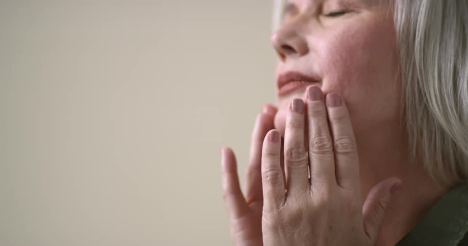 A beautifully aged gray-haired woman rubs her hands together as if there is lotion and then massages the wrinkles on her face as she closes her eyes.