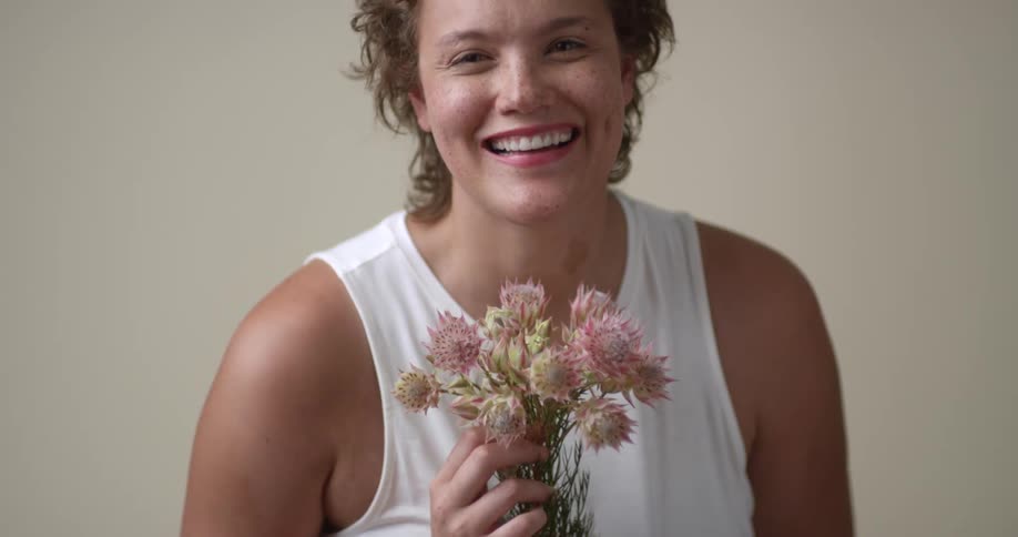 A young, white woman with short curly hair and freckles holds a bouquet of flowers and gives a dimpled smile to the camera, and then throws her head back and laughs.