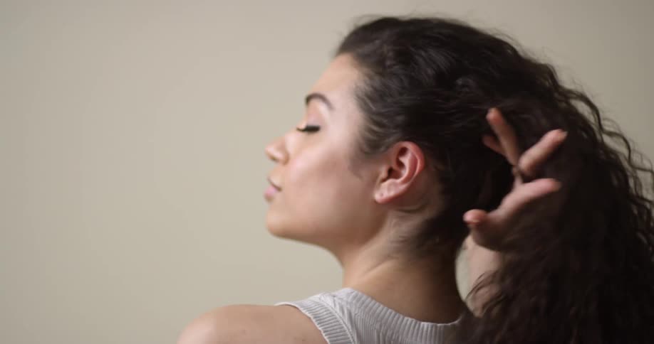 A young, female Pacific Islander with her back to the camera pulls her dark, curly hair up and over her shoulder and then turns to face the camera.