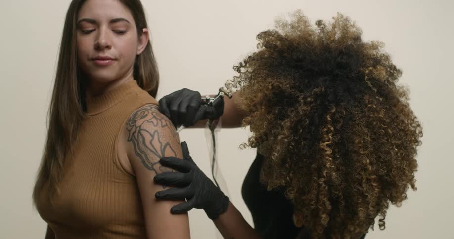 An ethnic, female tattoo artist with big, beautiful curly hair fills in a tattoo on the arm of a young caucasian woman with a tattoo gun.