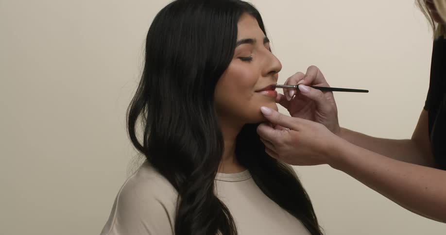 A young and ethnic model with dark, long hair is seated while a blonde, female makeup artist applies lipstick to her lips with a makeup brush.