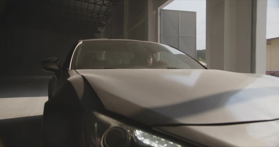 Man drives car through building with open doors showing another building and a hill in the background