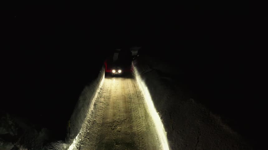 A vehicle travels along a road surrounded on either side by thick compacted snow at night