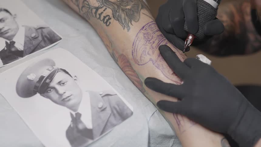 A tattoo artist is putting a tattoo of a person from a photo wearing military a unform on someones arm