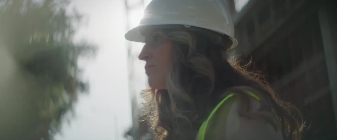 Woman wearing hardhat and vest stands near building under construction on sunny day