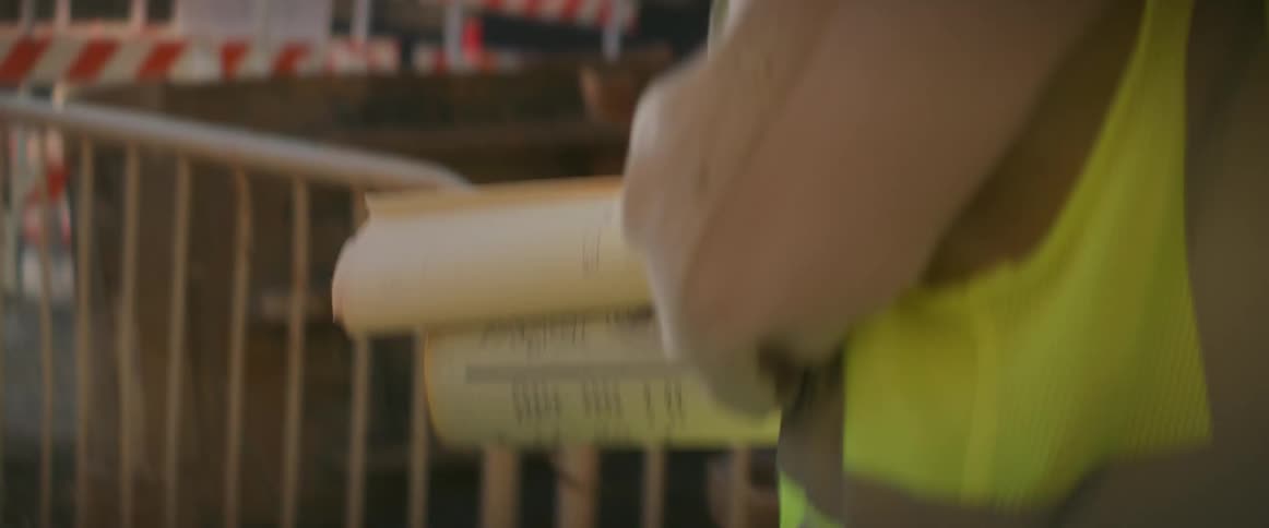 A female construction worker carries rolled up blueprints under her arm as she walks across a construction site in safety gear