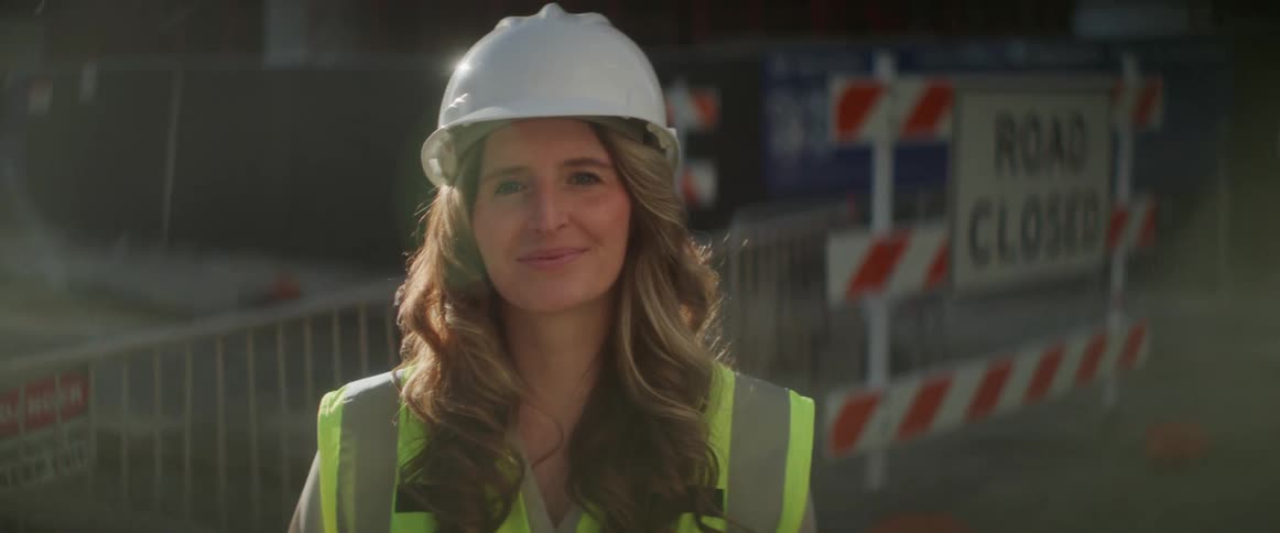A woman wearing a hardhat and reflective vest smiles standing in front of a construction site