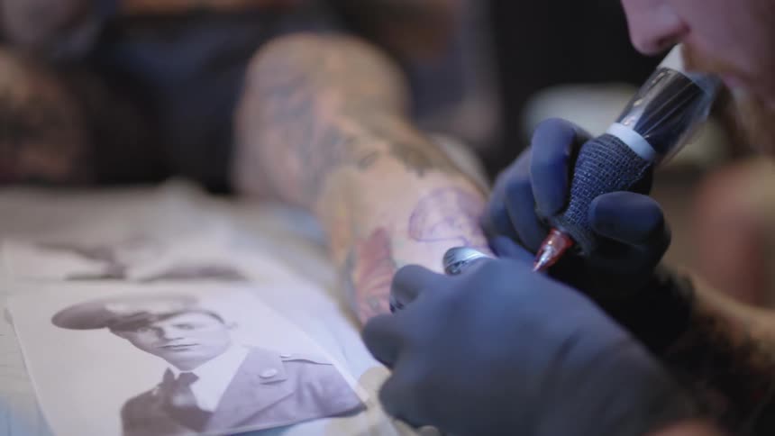 Tattoo artist adds tattoo to mans leg based on service man photo as client plays with mobile phone