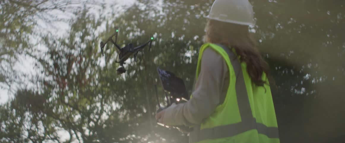 A female utility worker wearing a reflective vest is controlling a flying drone