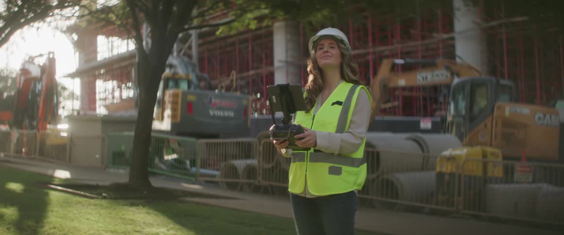 Female engineer with helmet and safety vest flies drone with remote controller in construction area