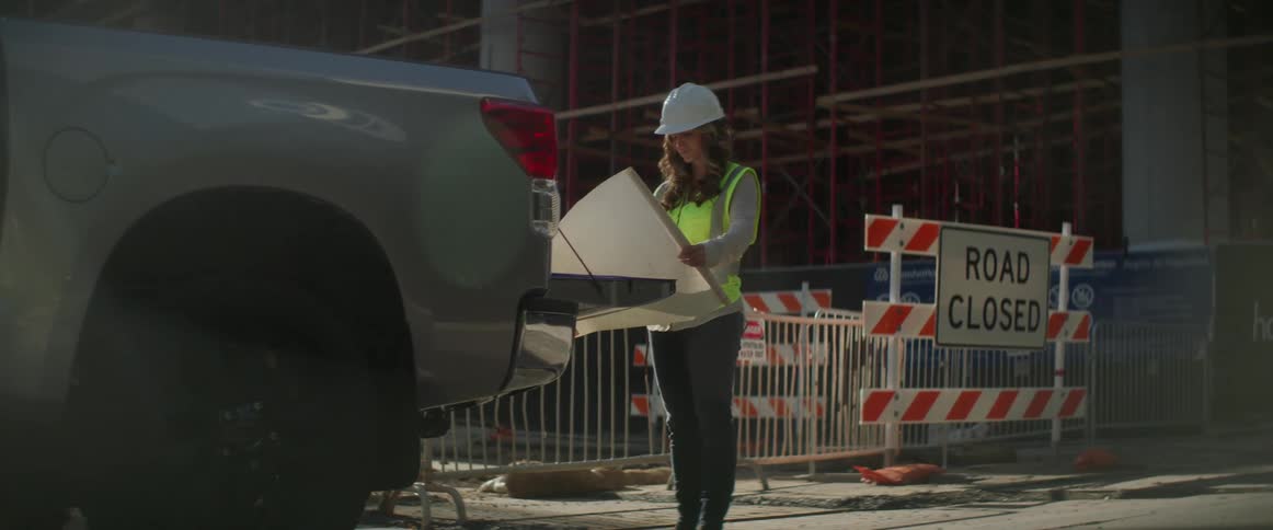 A woman with a hardhat and vest standing behind a pickup truck looking at a blueprint with a construction site in the background