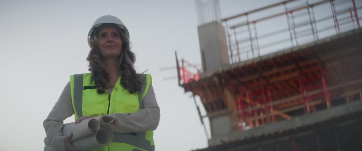 A woman with a hardhat and vest holding papers with the sky and clouds in the background