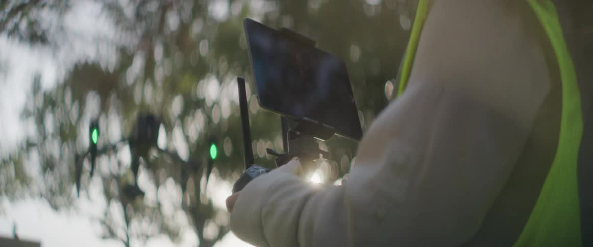 A woman with a hardhat and vest control in a drone with trees the sky and clouds in the background