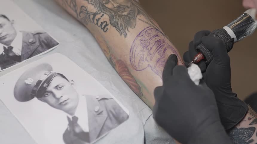 Male tattoo artist with black gloves applies tattoo on mans arm by looking at photogrph in the studio