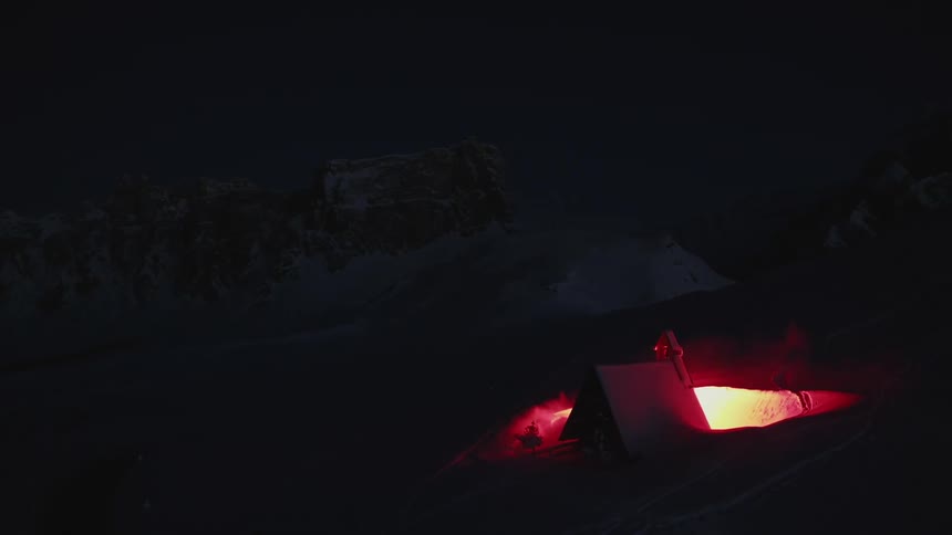 Aerial view of a small cabin in the snow illuminated by a red flare at night