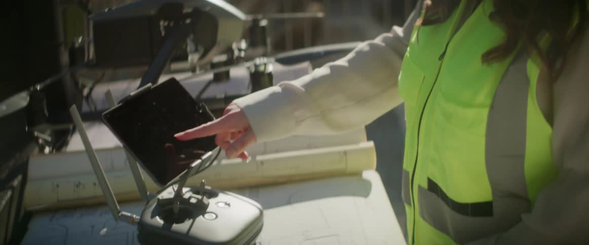 Female engineer with white helmet brown wavy hair and safety vest uses remote controller to control flying drone in the construction area