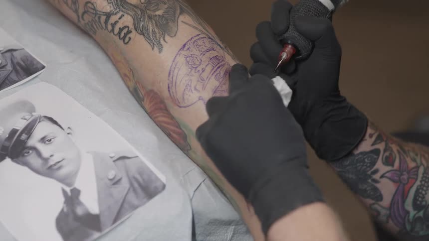 A tattoo artist references a photo while tattooing the arm of a customer