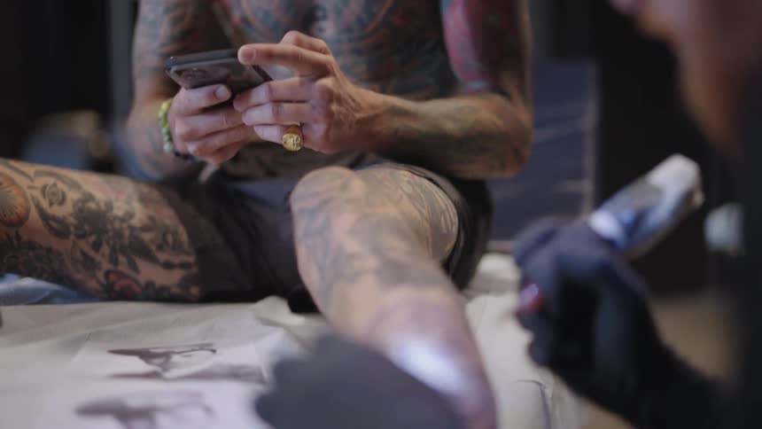 Man texting on the phone while a tattoo artist works on his leg