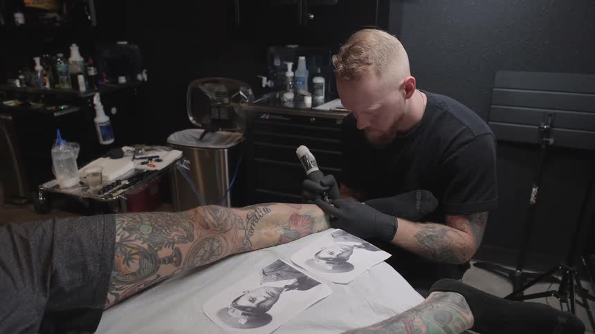 A tattoo artist tattoos a customers arm while referencing a photograph of a soldier