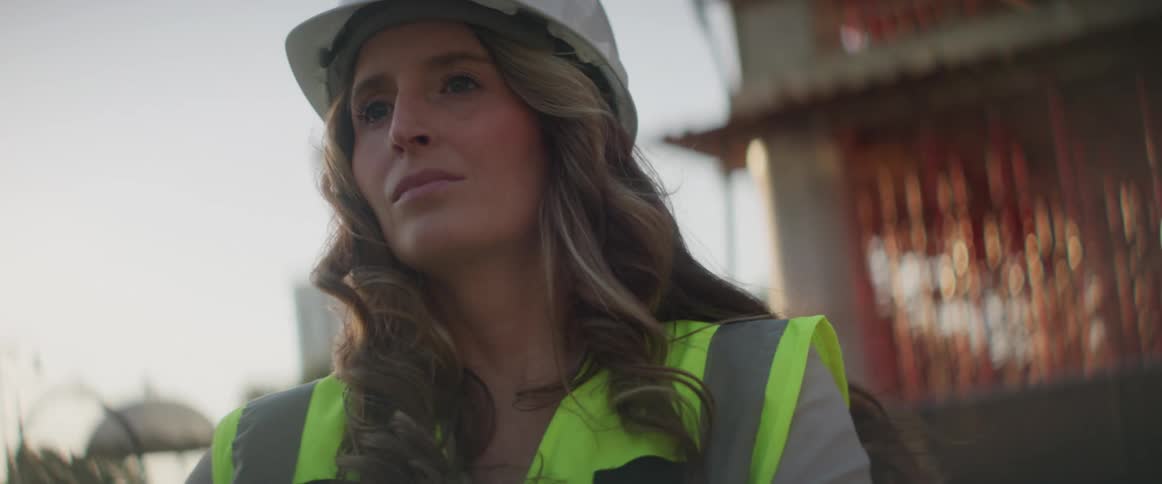 A female construction worker is wearing a hard hat and looking around