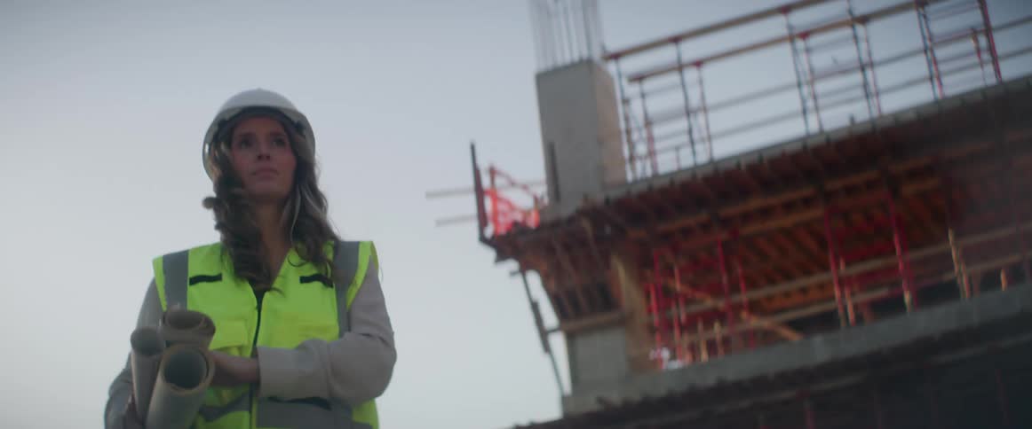 A woman wearing a hardhat and reflective vest walks onto a construction site carrying rolled up plans
