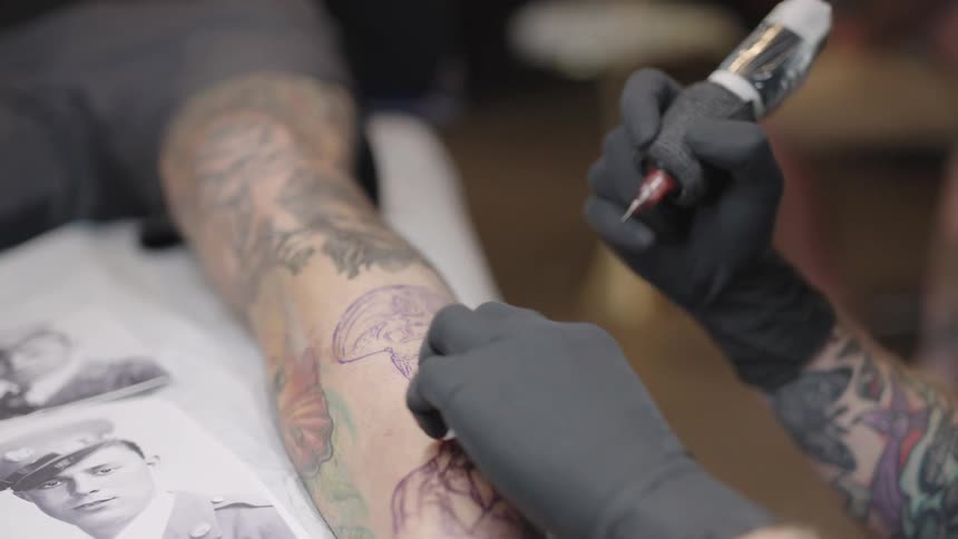 A tattoo artist pauses tattooing to dab at a clients arm with gauze