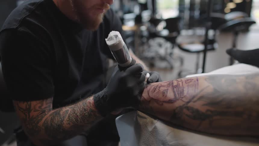 Tattoo artist with beard black shirt and gloves applies tattoo on mans arm covered with tattoos in the studio