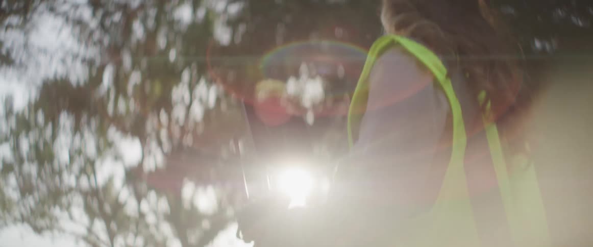 A woman in a safety vest and hard hat operates a remote controlled drone outdoors during the day