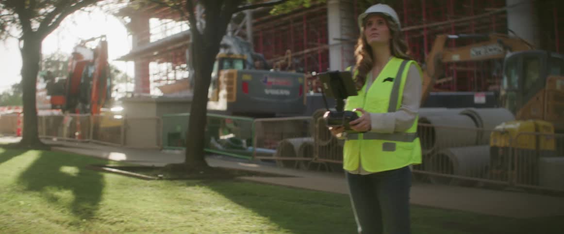 A woman with a hardhat and vest controlling a drone with a construction site trees the sky and clouds in the background
