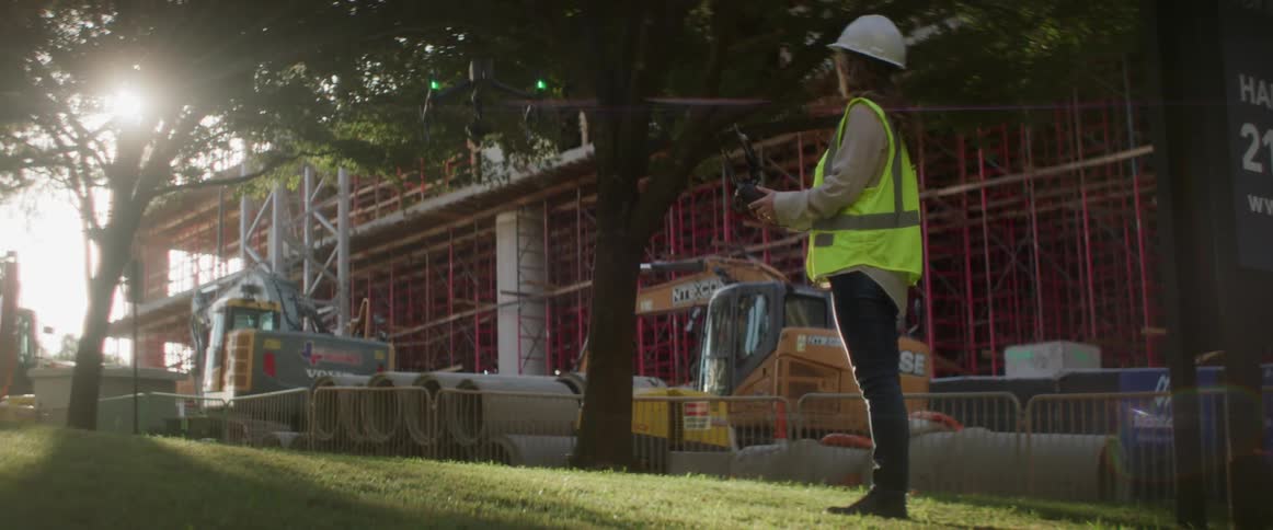 A woman with a hardhat and vest control in a drone with trees the sky and clouds in the background