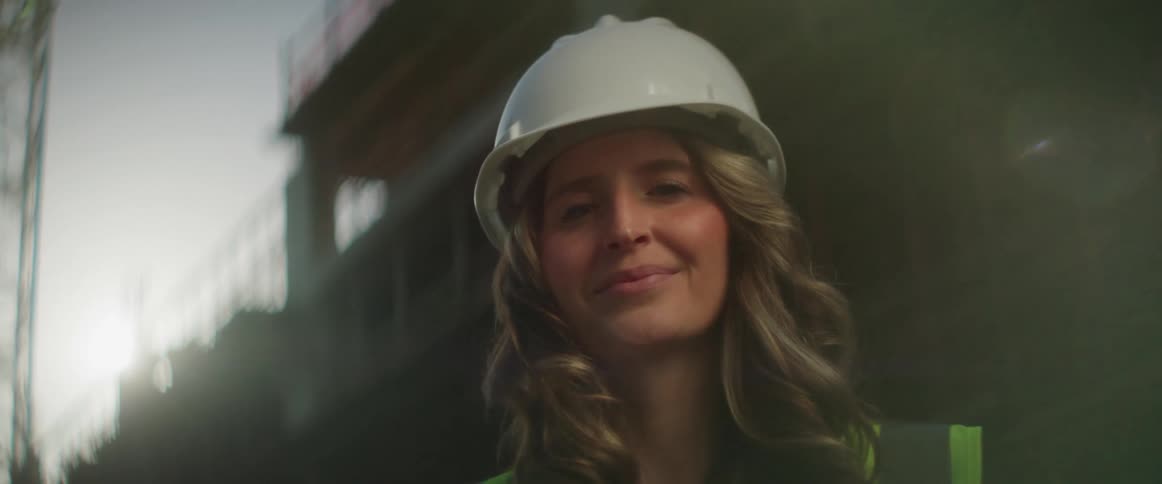 A woman with long hair wearing a hardhat and reflective vest stands with a construction site behind her