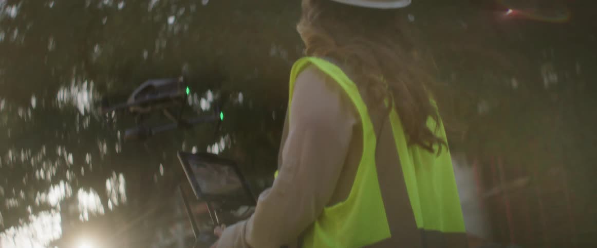 A woman in hardhat and reflective vest controls a drone near a construction site