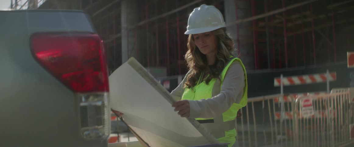 Female engineer with white helmet brown wavy hair and safety vest looks at map in construction area