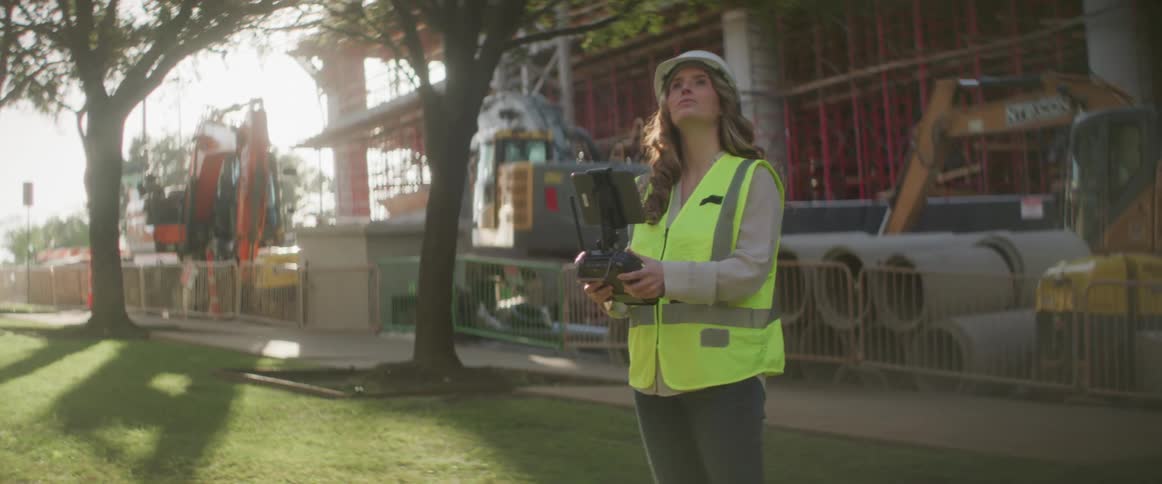A female construction worker is wearing a hard hat and operating a drone