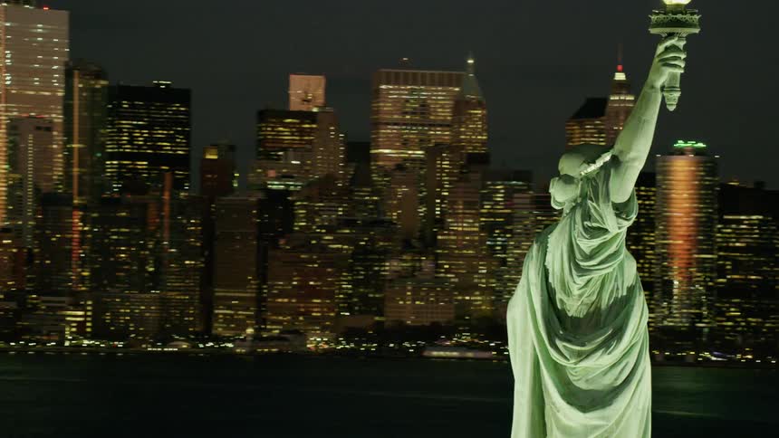 Statue of liberty stands across harbor from downtown and a suspension bridge both lit by lights at night