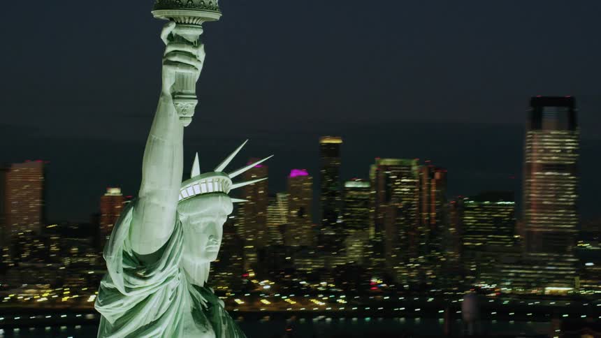 The statue of liberty holds her torch high in the air with tall city building lit up in the distance