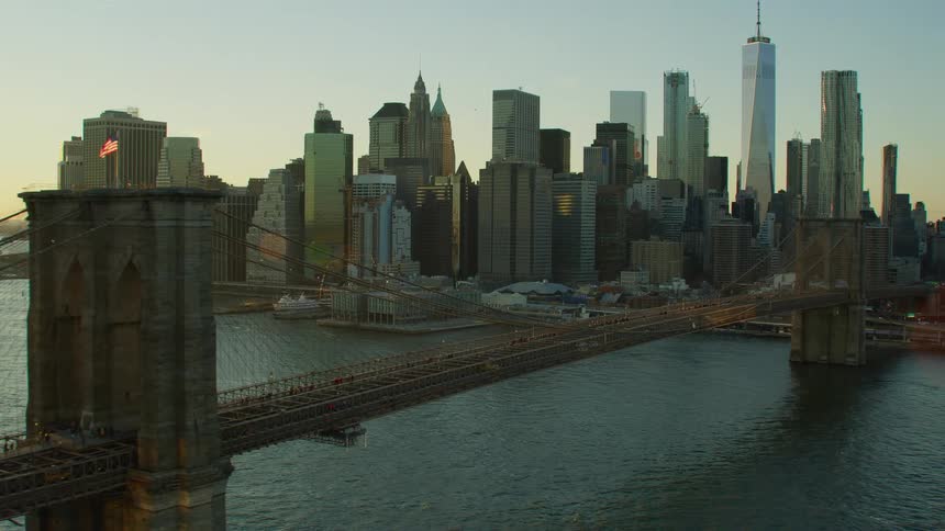Cars travel across a bridge over a river to a big city with tall skyscrapers high in a clear blue sky