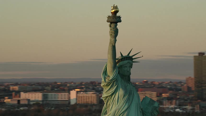 The statue of liberty holds her torch high in the air with tall city buildings in the distance