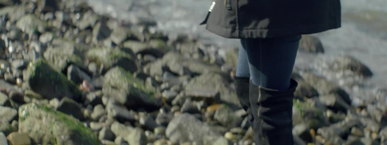 A woman walks on the rocks by the water near a city