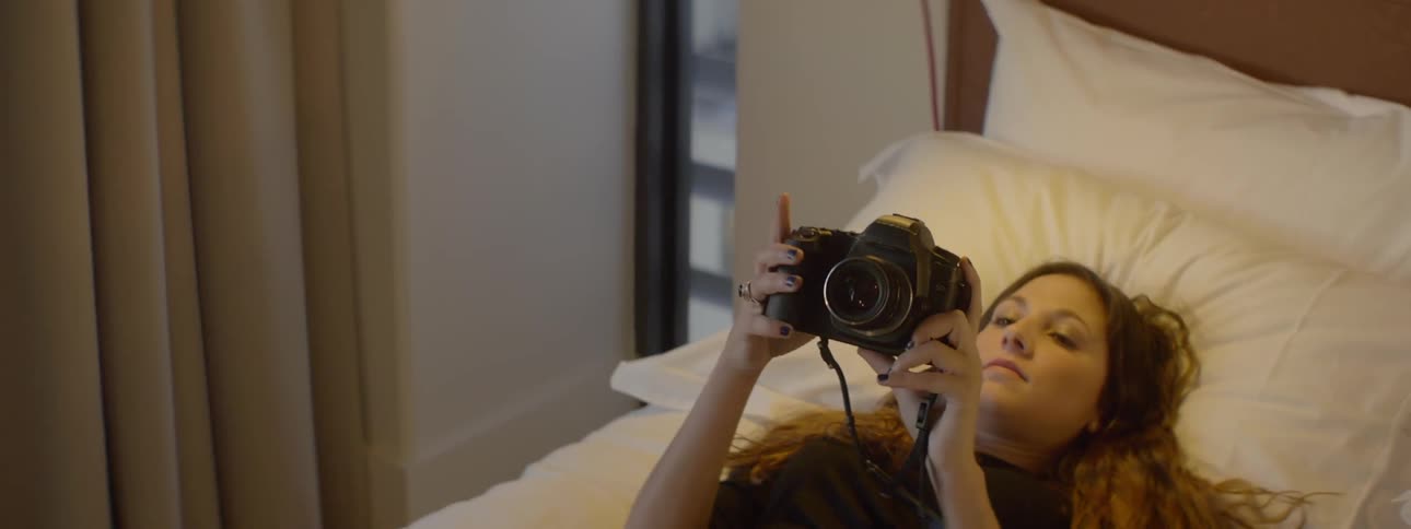 A woman lies down on a bed and plays with a camera