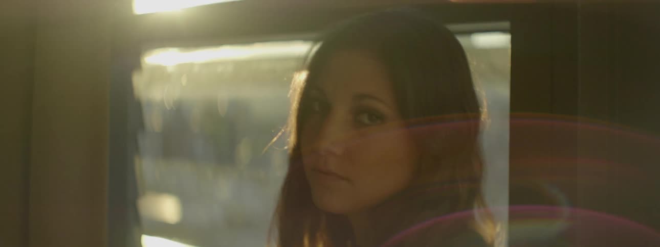 Woman looking out a window at sunset and then turning to look into the camera