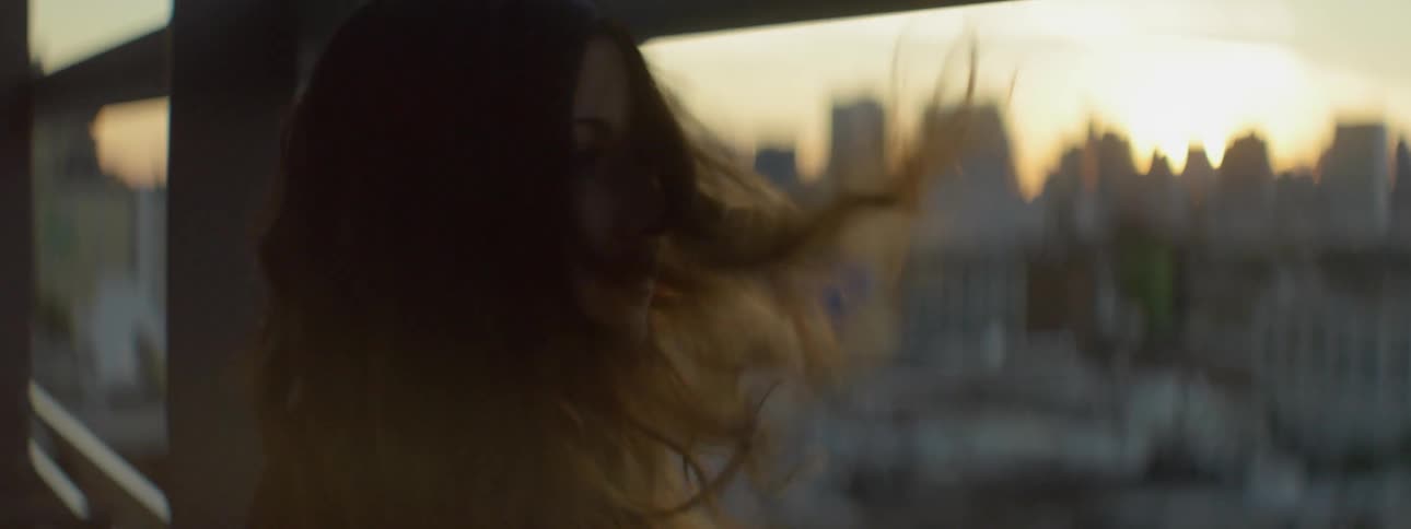 A woman standing by a window looks outside as wind blows her hair